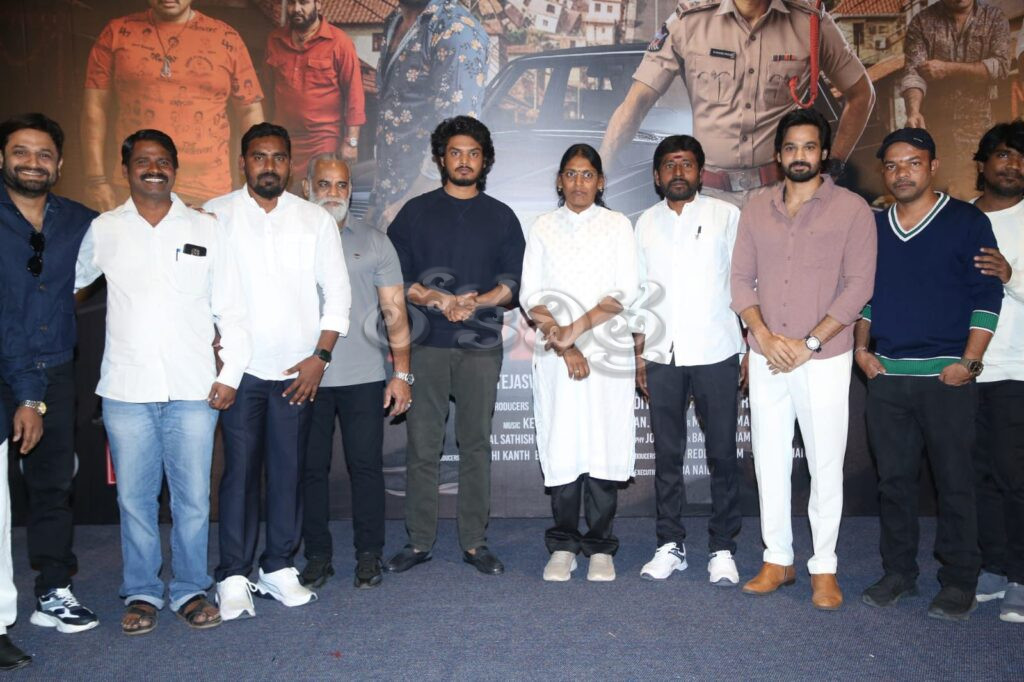 Group of individuals posing together at a promotional event for an Indian film, with a backdrop featuring movie characters.